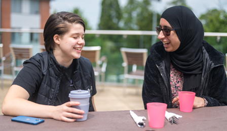 two female students sitting at a table outside holding coffee cups looking towards each other and smiling