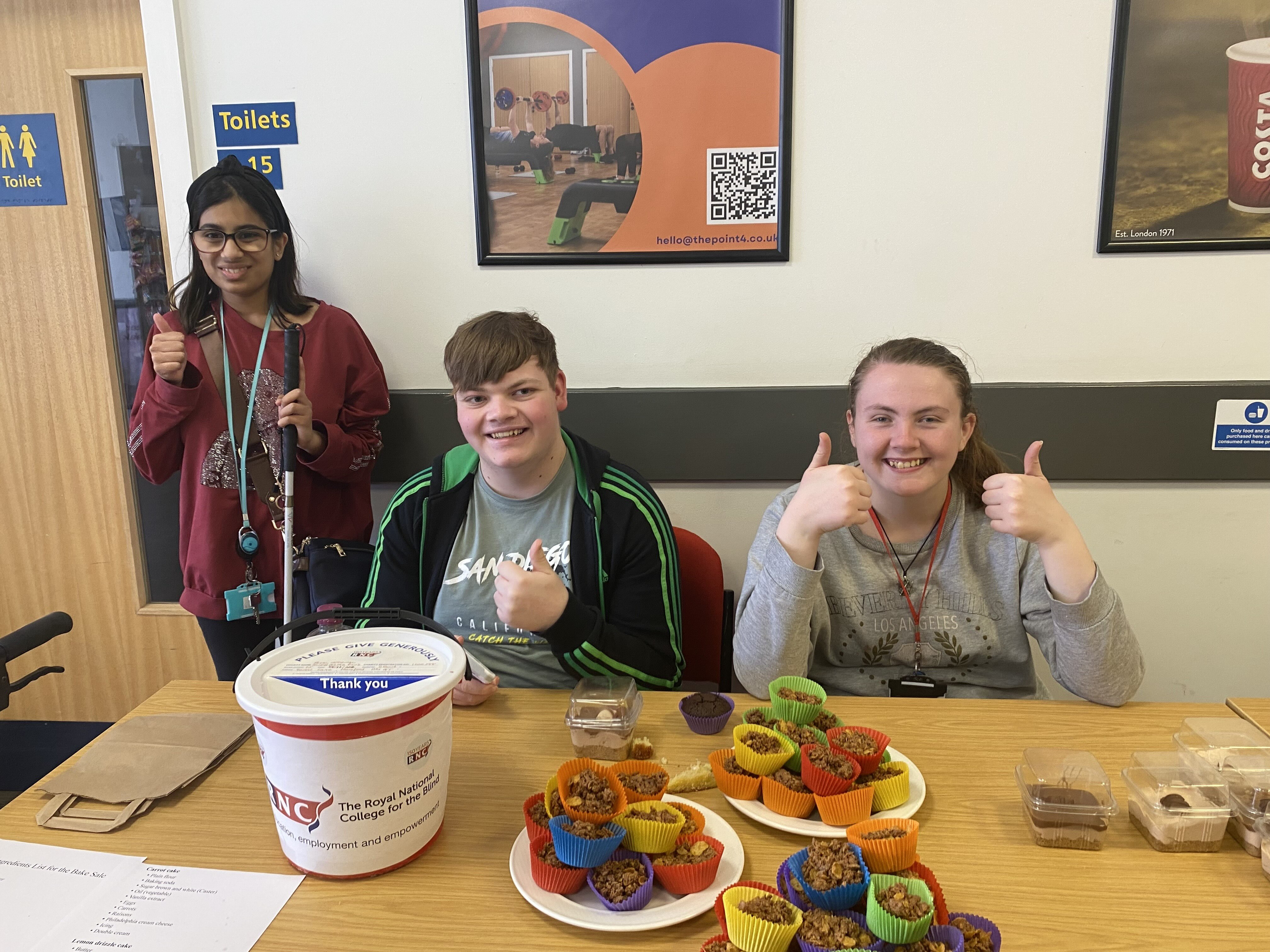 2 sit and 1 person stands behind a table with the cakes displayed in front of them - they all have big smiles