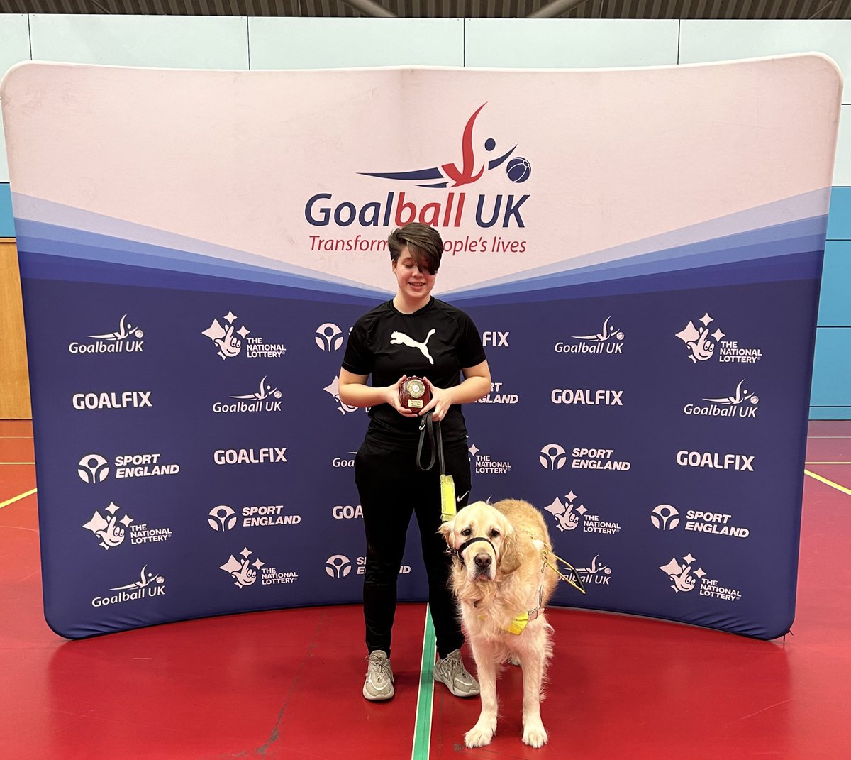 Freya and guide dog Lennon stand in front of the Goalball UK banner with her award