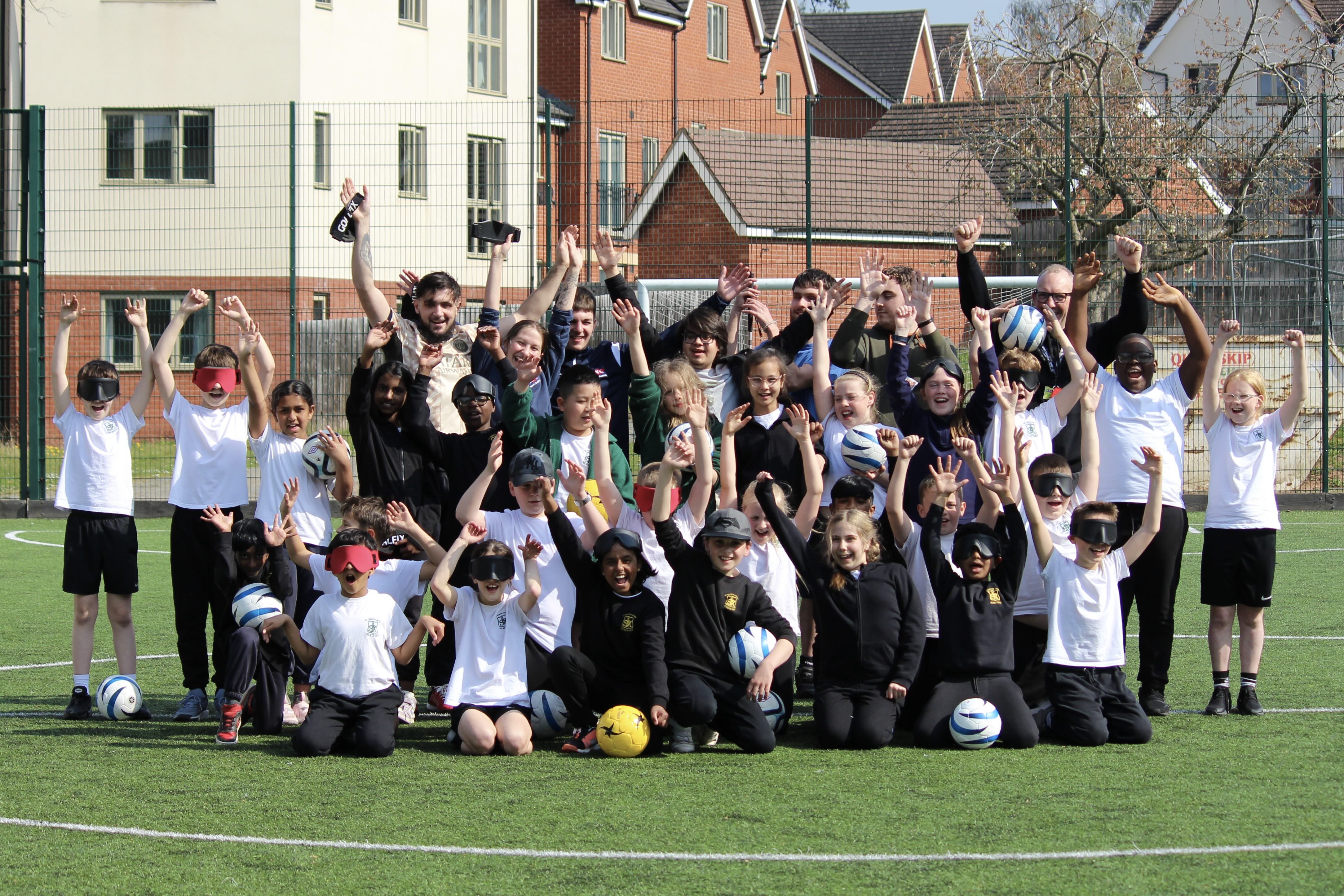 The group stand in the centre of the football pitch some kneeling, some standing, some wearing or holding eye shades and footballs