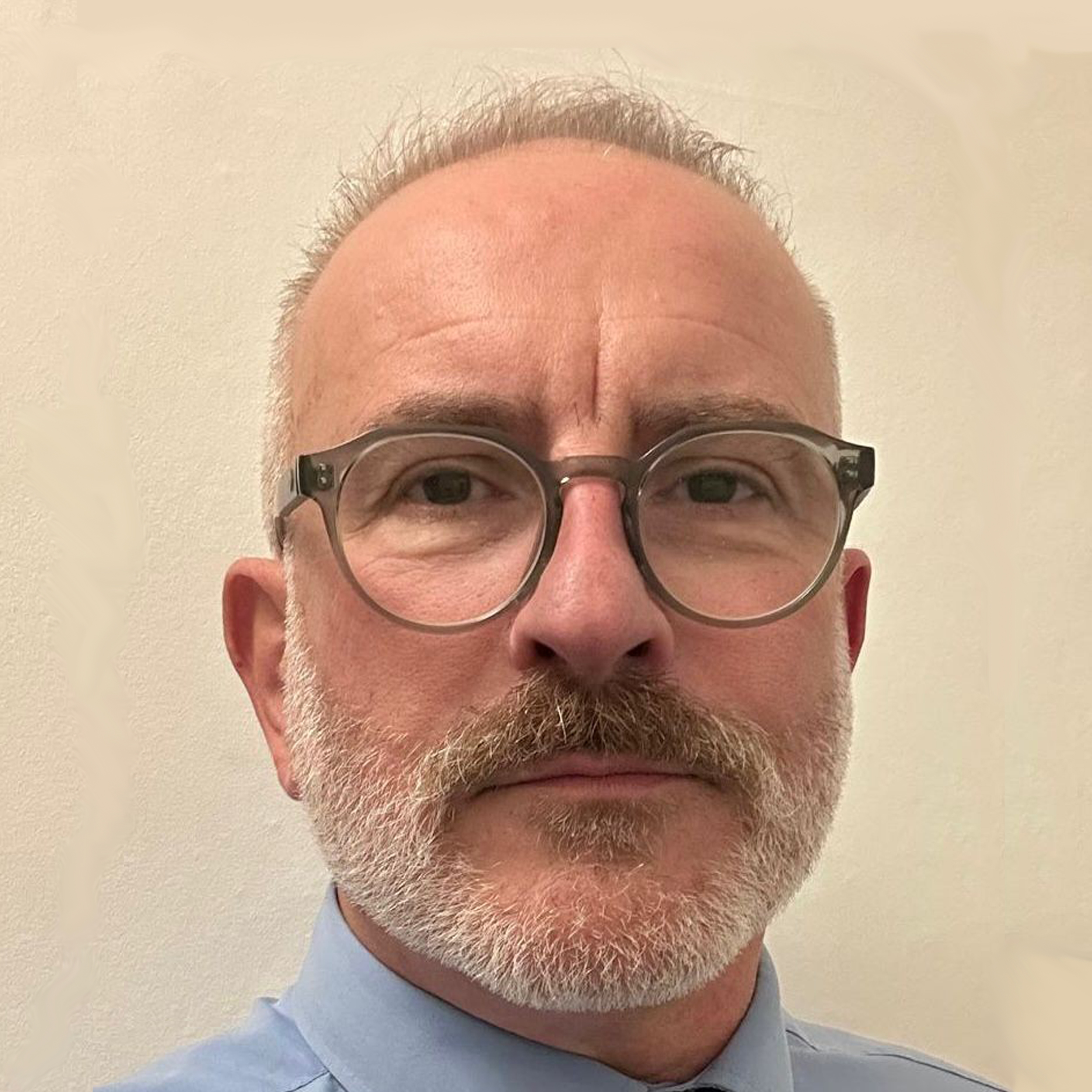 a head and shoulders picture of a male with gray spikey hair and gray facial hair - he is wearing glasses