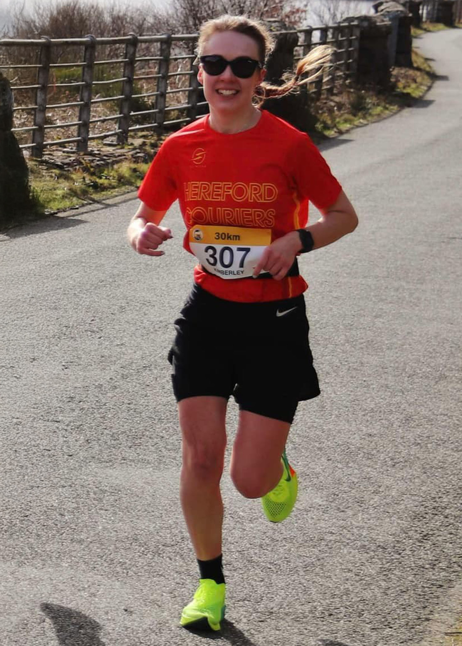 Kim running on a road with a fence behind her she is wearing a red Hereford Couriers running top and black shorts