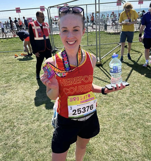 Kim in a red couriers vest holding her marathon medal she has a broad smile and is holding a bottle of water and phone in her left hand