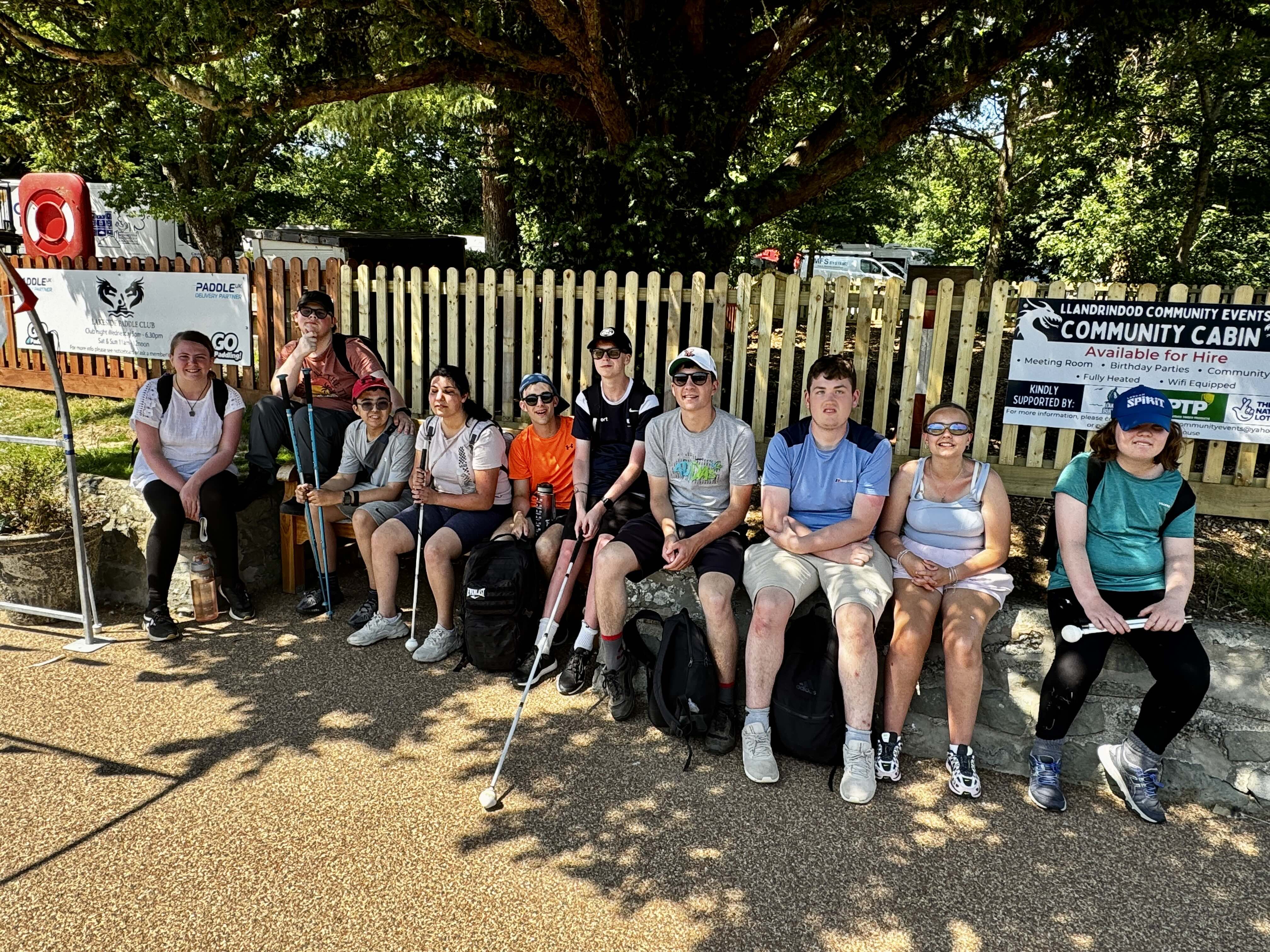 10 students sitting on a low wall under a tree for shade on a hot sunny day