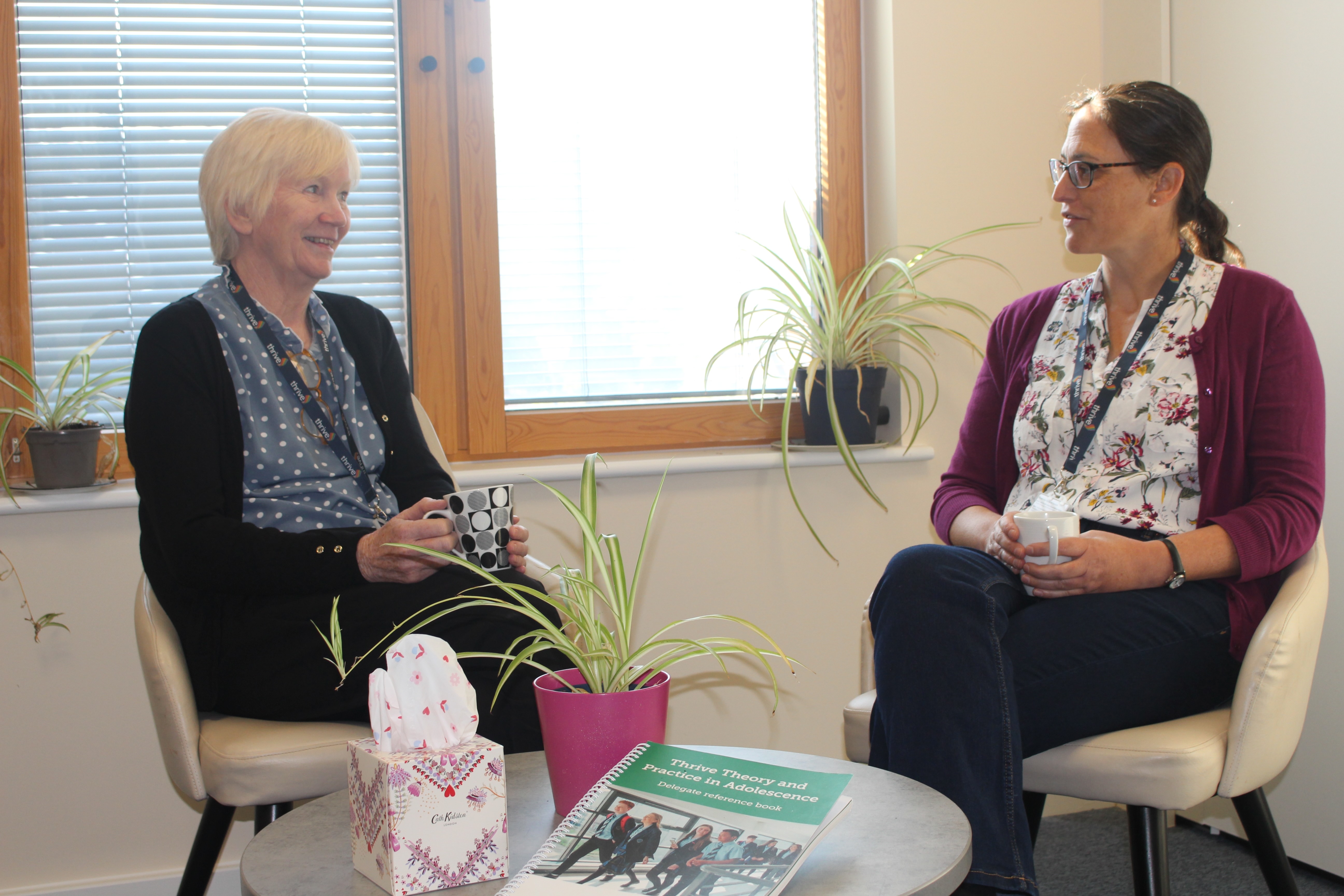 two people sitting facing each other and talking -they hold mugs and there is a coffee table in front of them with a spider plant on it, a box of tissues and a book titled 'Thrive Theory and Practice in Adolescence'