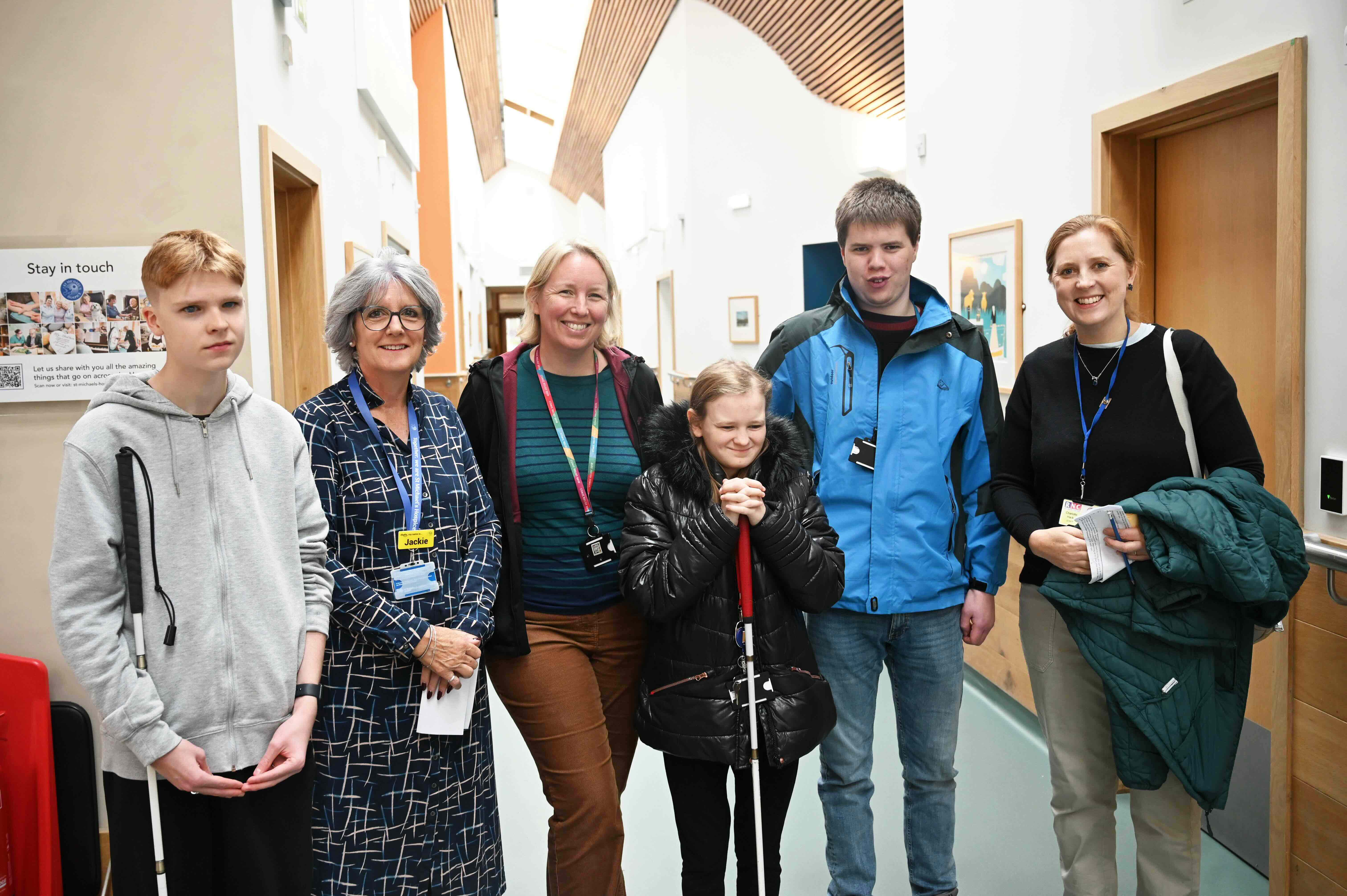 a group of six people stand in a corridor - two of them holding canes - they are all smiling for a photograph