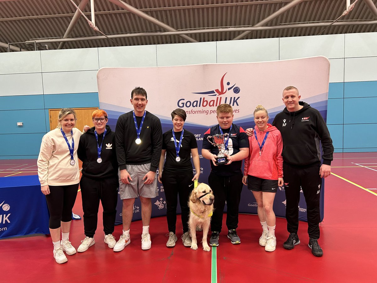 The team with a guide dog, guides and coaches stand in front of a Goalball UK stand wearing their gold medals