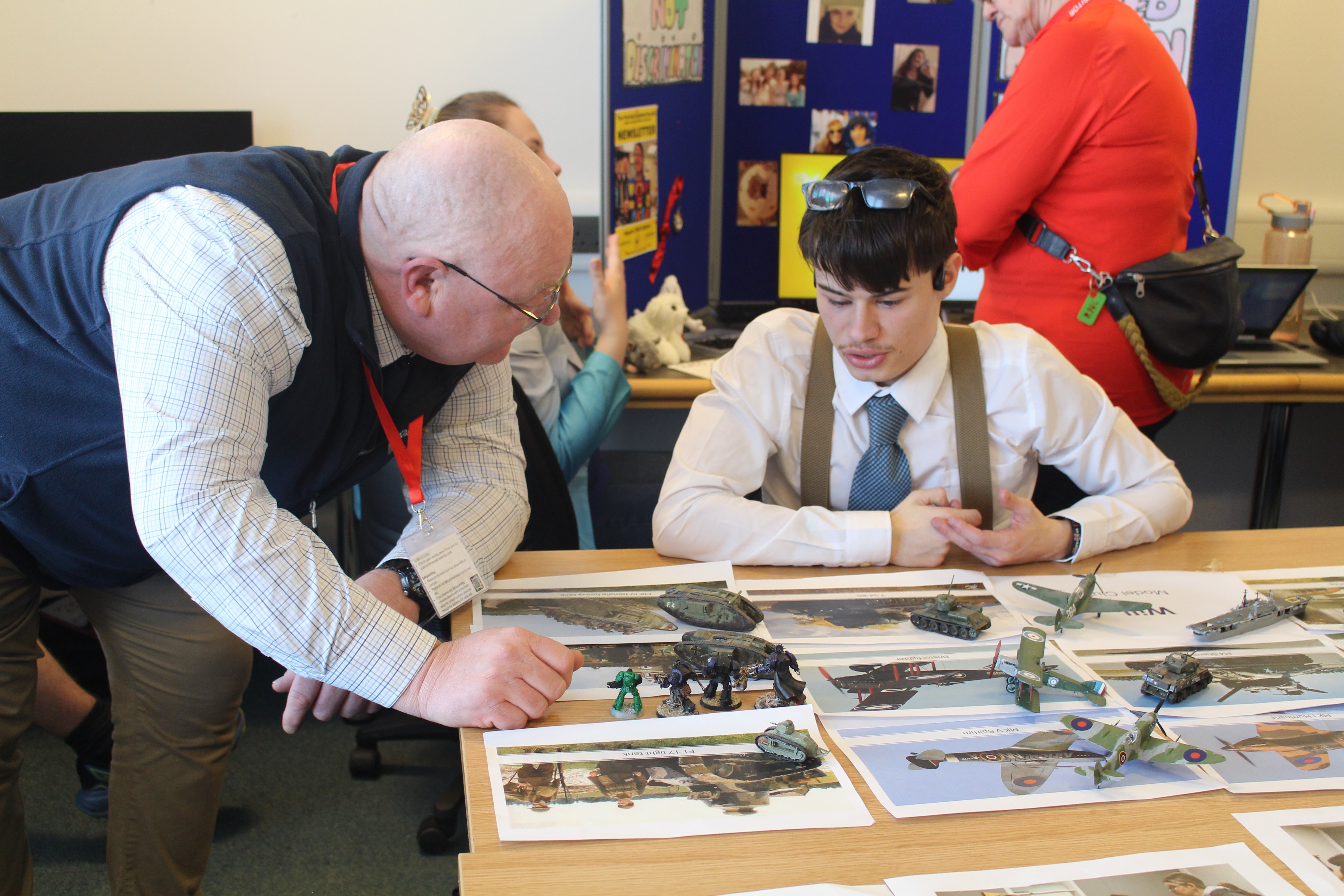 a visitor leaning on the table discusses with a student the models of planes and tanks displayed on the table
