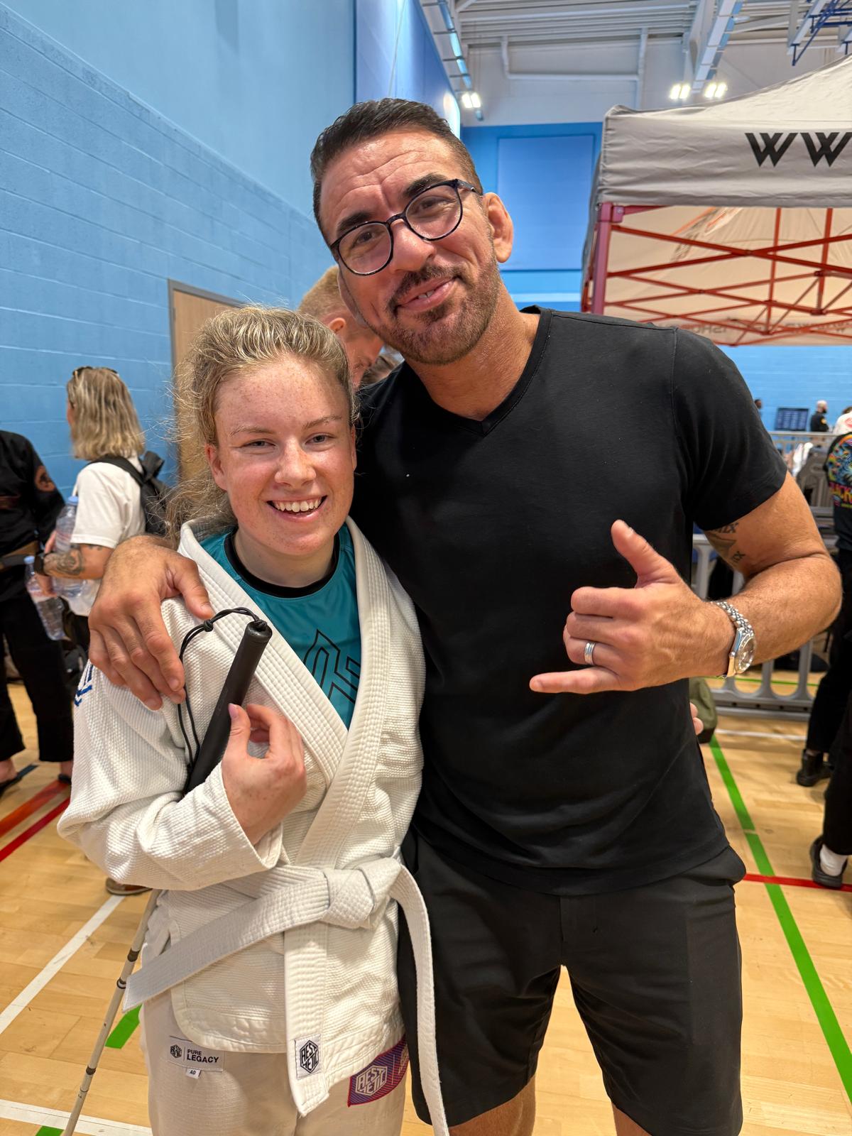 a competitor dressed in white gi and holding a cane, is next to a man dressed in black t-shirt and trousers