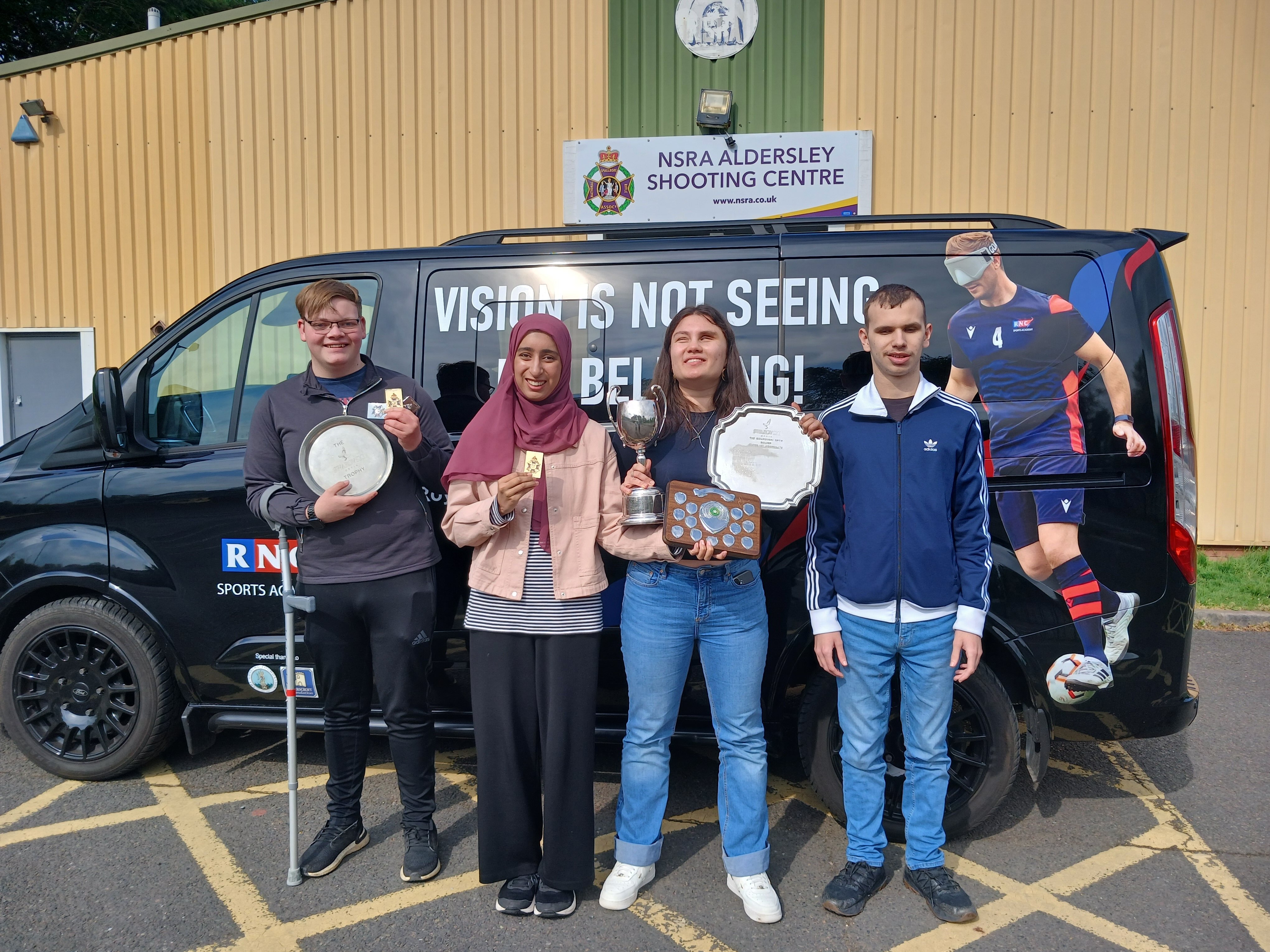 four students standing in front of a small black van holding up their trophies and smiling for the camera