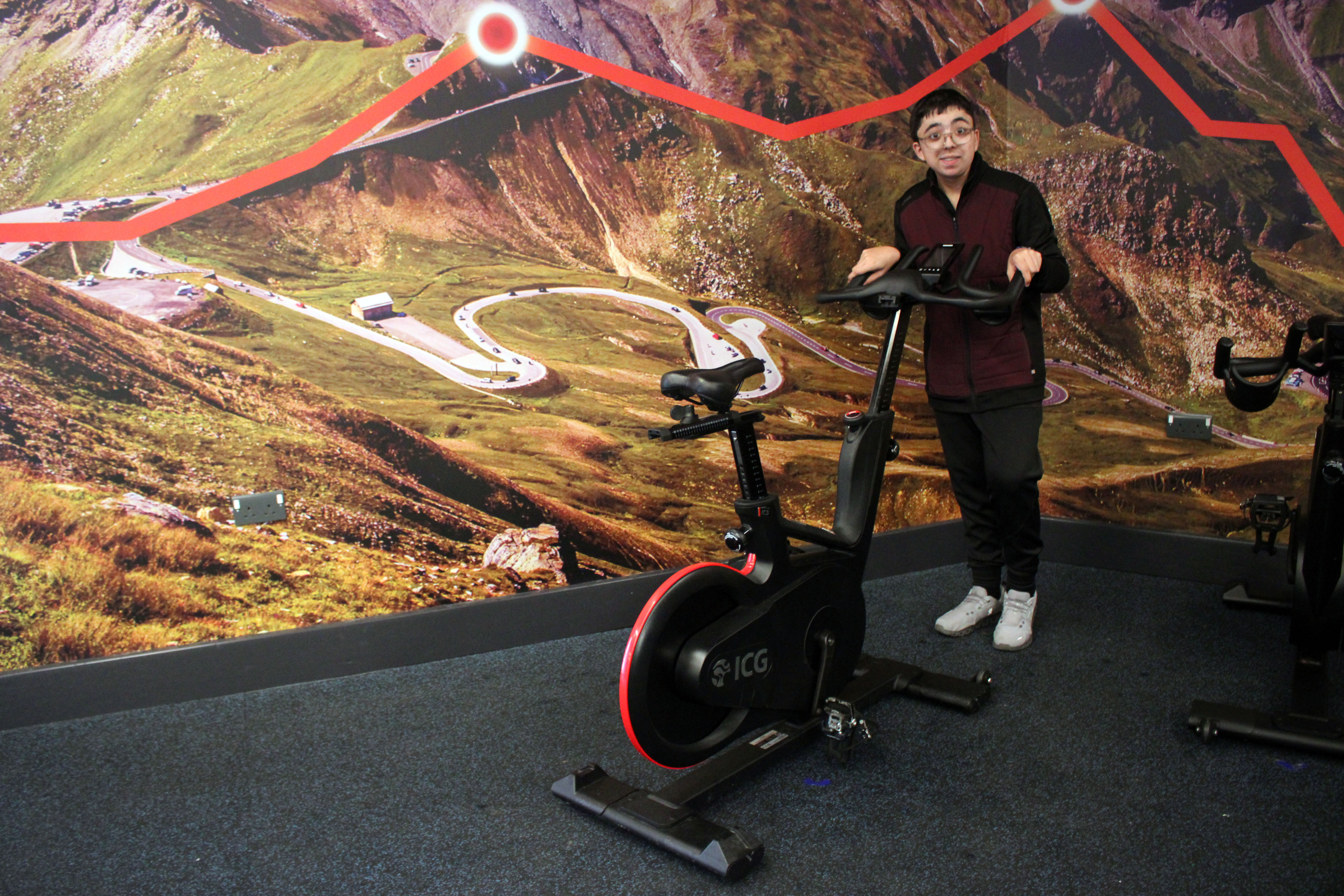 Emilio in the spin studio there's a giant mural behind him of a cycle route going across mountains. Emilio is putting a spin bike. away by pulling it on castors