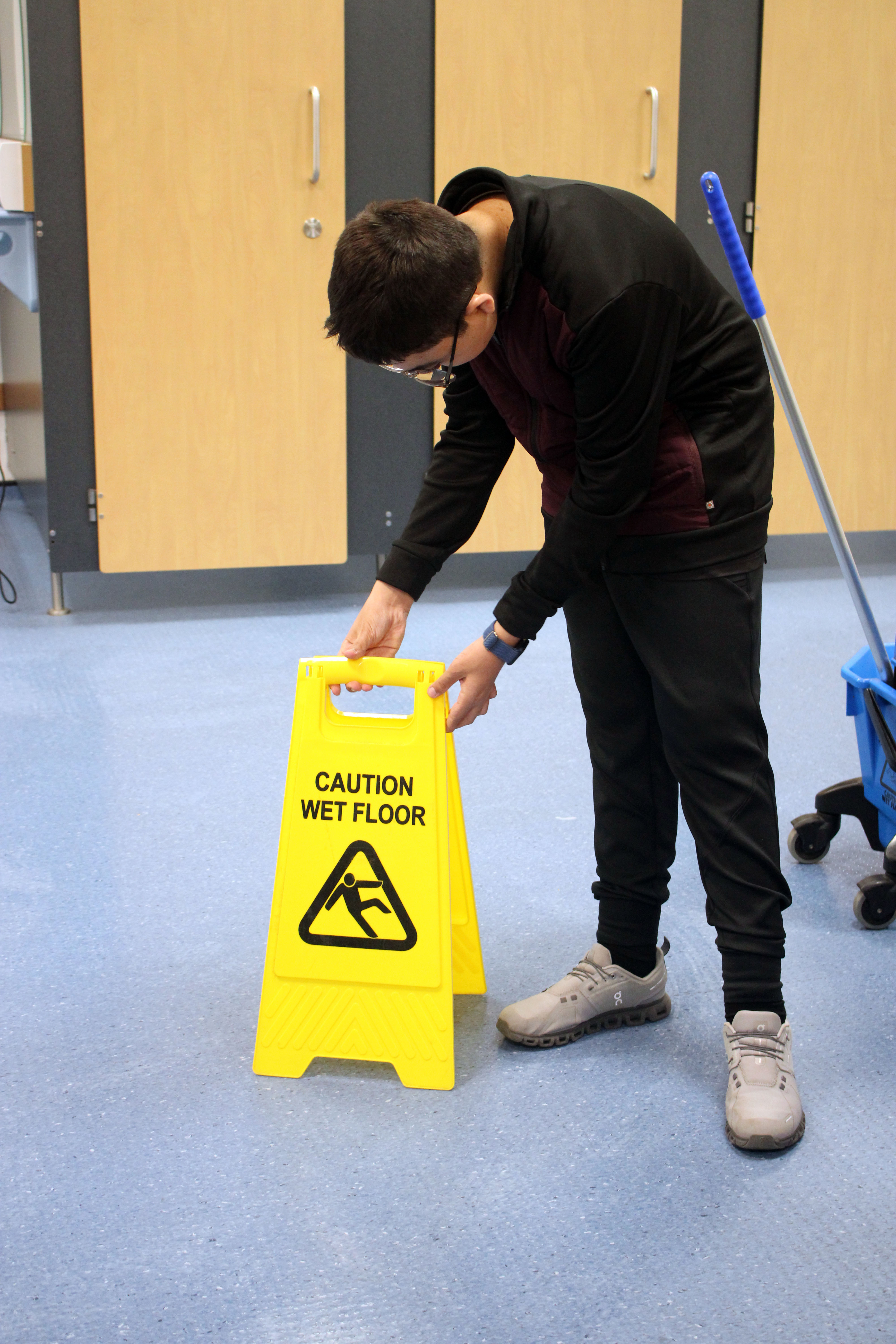 Emilio stands next to a big blue mop bucket with the mop standing inside it. He is putting out a bright yellow 'wet floor' sign